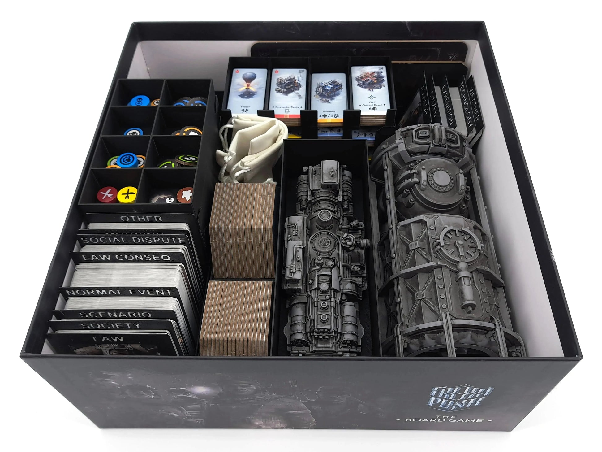 The Frostpunk board game box partially packed, showing card dividers on the left, a token tray with colorful circular tokens, and several large cardboard player boards and maps stacked on the right.