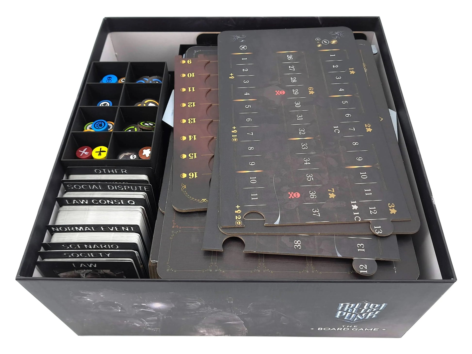 The Frostpunk board game box partially packed, showing card dividers on the left, a token tray with colorful circular tokens, and several large cardboard player boards and maps stacked on the right.