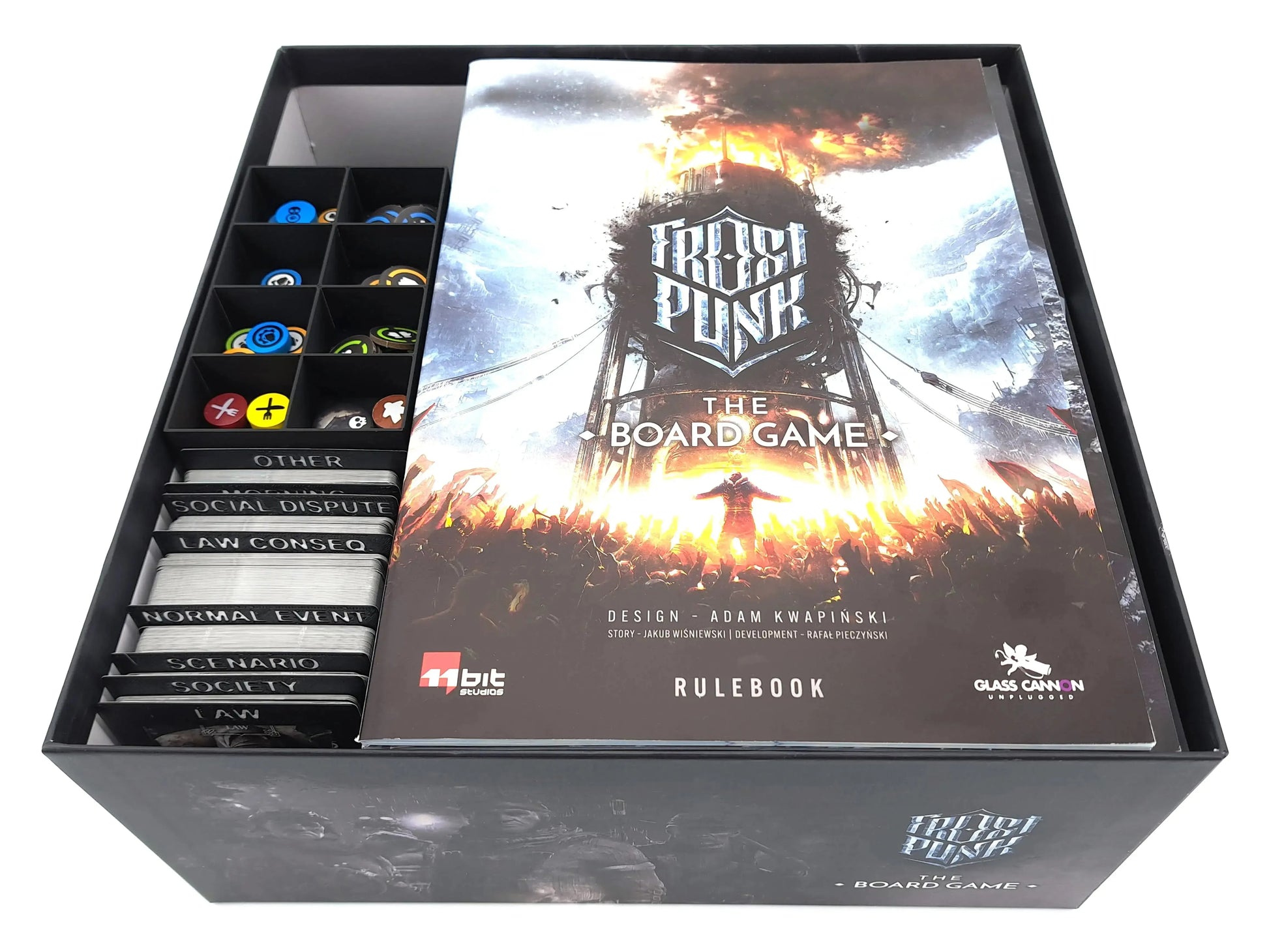 The final stage of packing the Frostpunk board game box. The card and token organizers are visible on the left, while the official rulebook rests flat on top of the other components, filling the box to the brim.