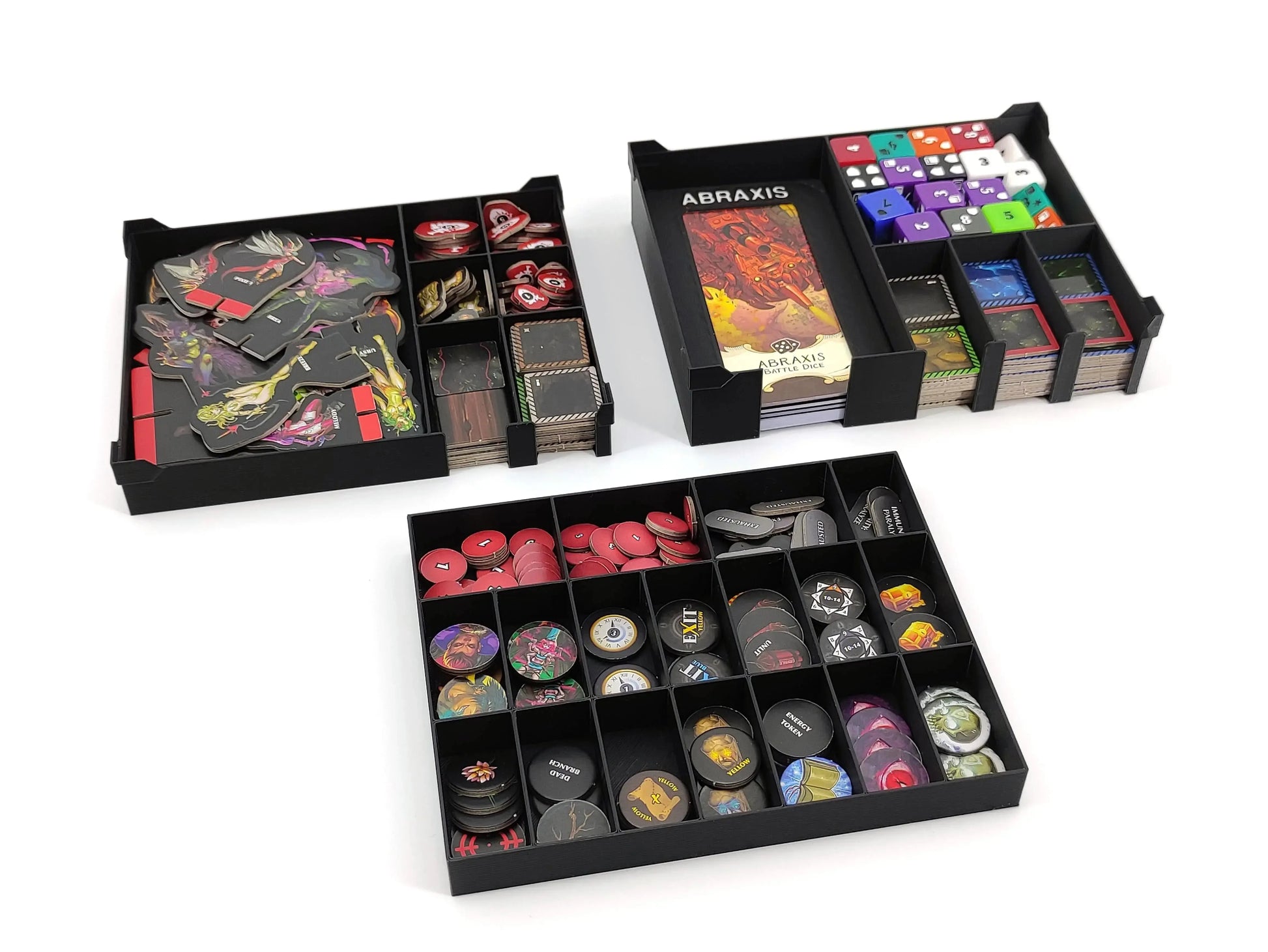 Three modular black trays for Middara board game components, organized with colorful dice, cardboard tokens, and small circular game markers.
