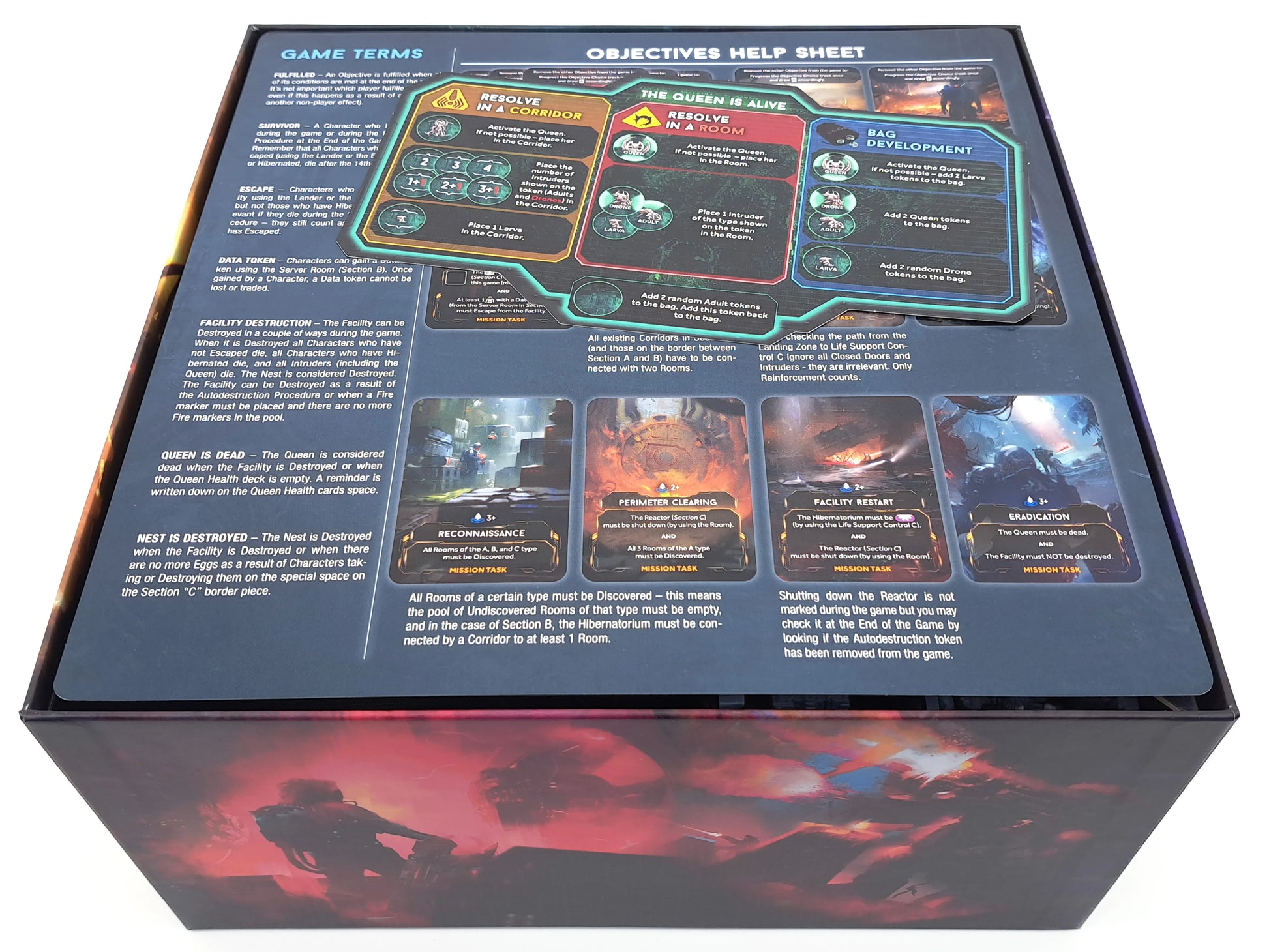Oberseite der Nemesis-Retaliation-Spielbox, auf der ein farbiges Objectives Help Sheet und die Game Terms Übersicht liegen. Zu sehen sind Regeln, Icons, Missionskarten und grafische Darstellungen der Spielmechaniken. Die Box zeigt das typische Awaken-Realms-Artwork.