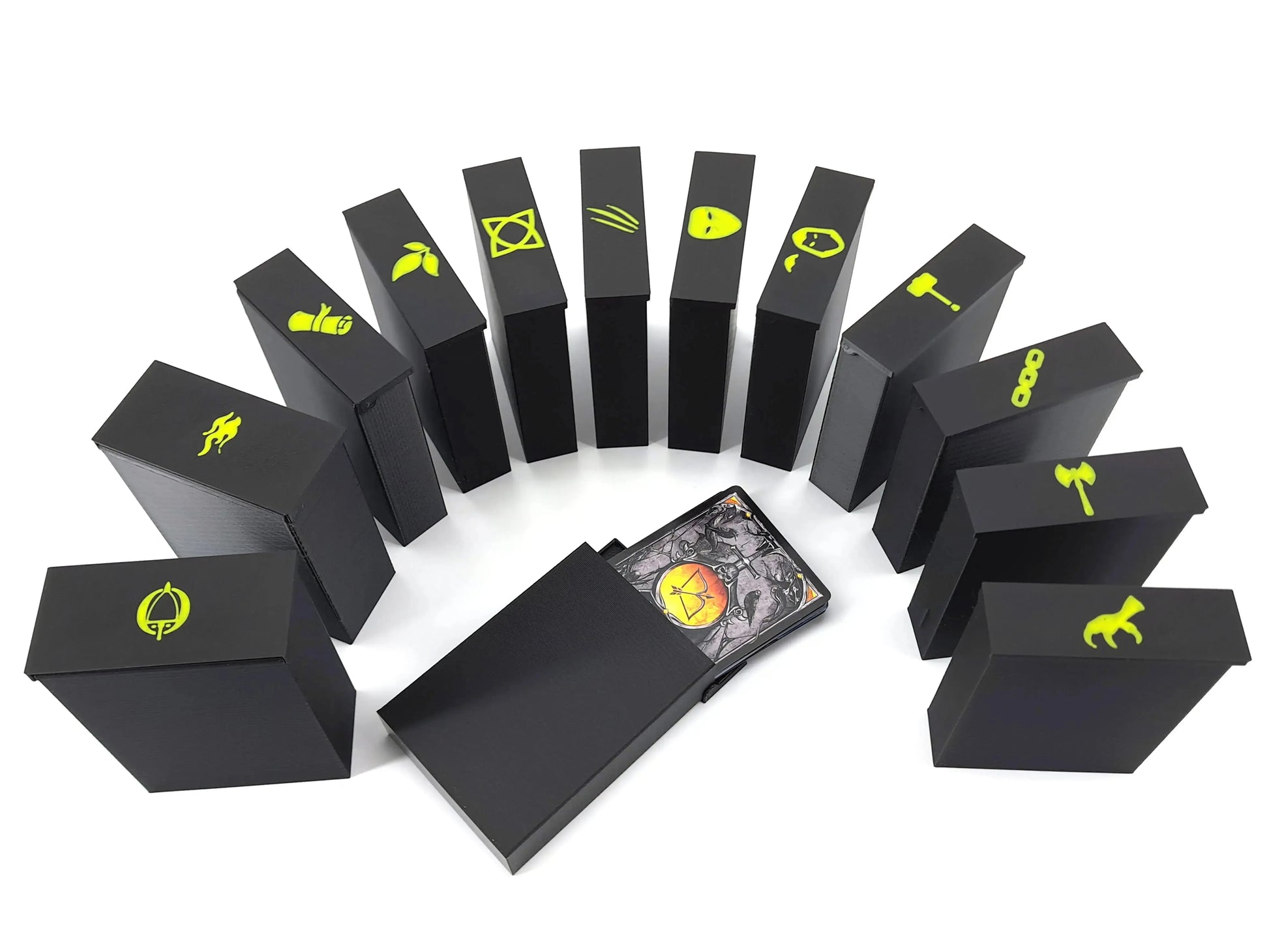 Photo of multiple black 3D-printed card-storage boxes arranged in a semicircle. Each box features a yellow symbol on top representing different game components. One box in the center is pulled open, revealing a stack of illustrated game cards inside.