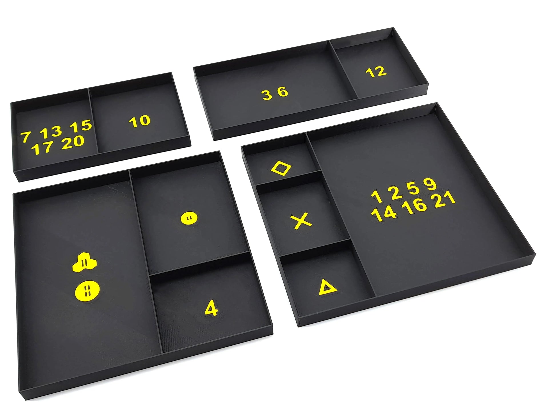 An arrangement of four black organizer inserts for a board game, featuring various yellow numbers and symbols in the compartments. The numbers include 7, 10, 12, 13, 14, 15, 16, 17, 20, 21, 36, 1259, 4, and various symbols such as an "X", a triangle, and a diamond symbol. The inserts are used for the organized storage of game components.